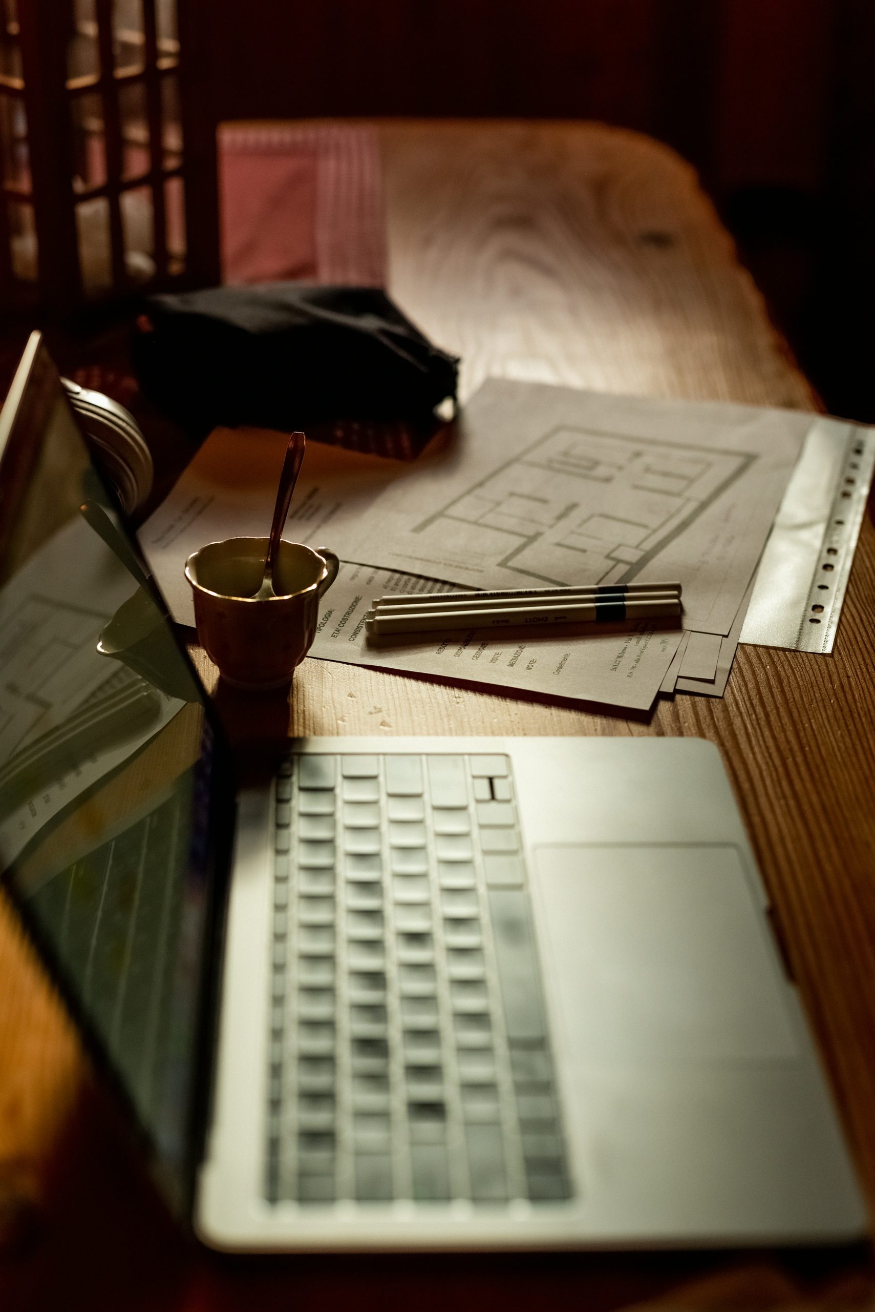 Office desk with a laptop showing a comparative chart of software features, surrounded by construction blueprints and a coffee cup.