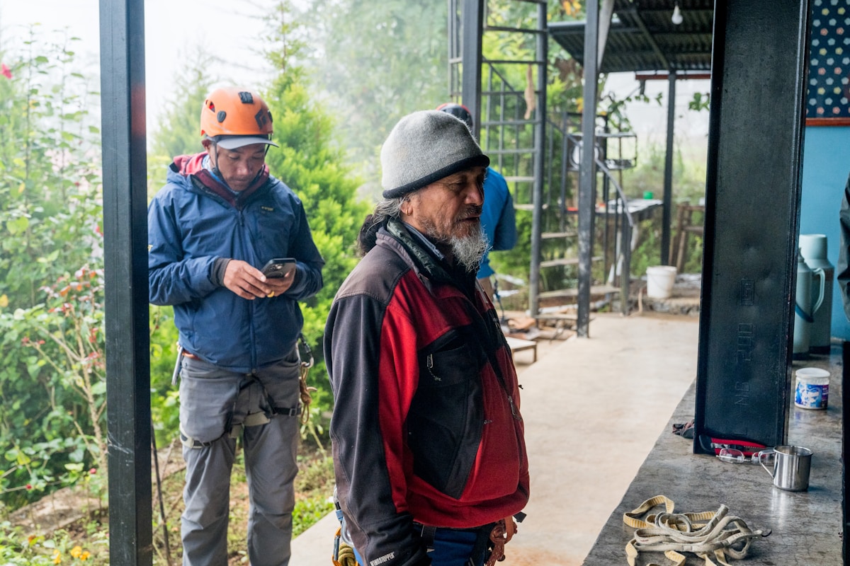 Deux hommes en tenue de travail, l'un consultant son téléphone, l'autre debout dans un environnement extérieur verdoyant.